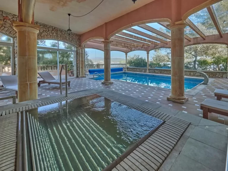 Área cubierta con piscina rectangular y jacuzzi con estructura de vigas de madera y ventanales en Mallorca