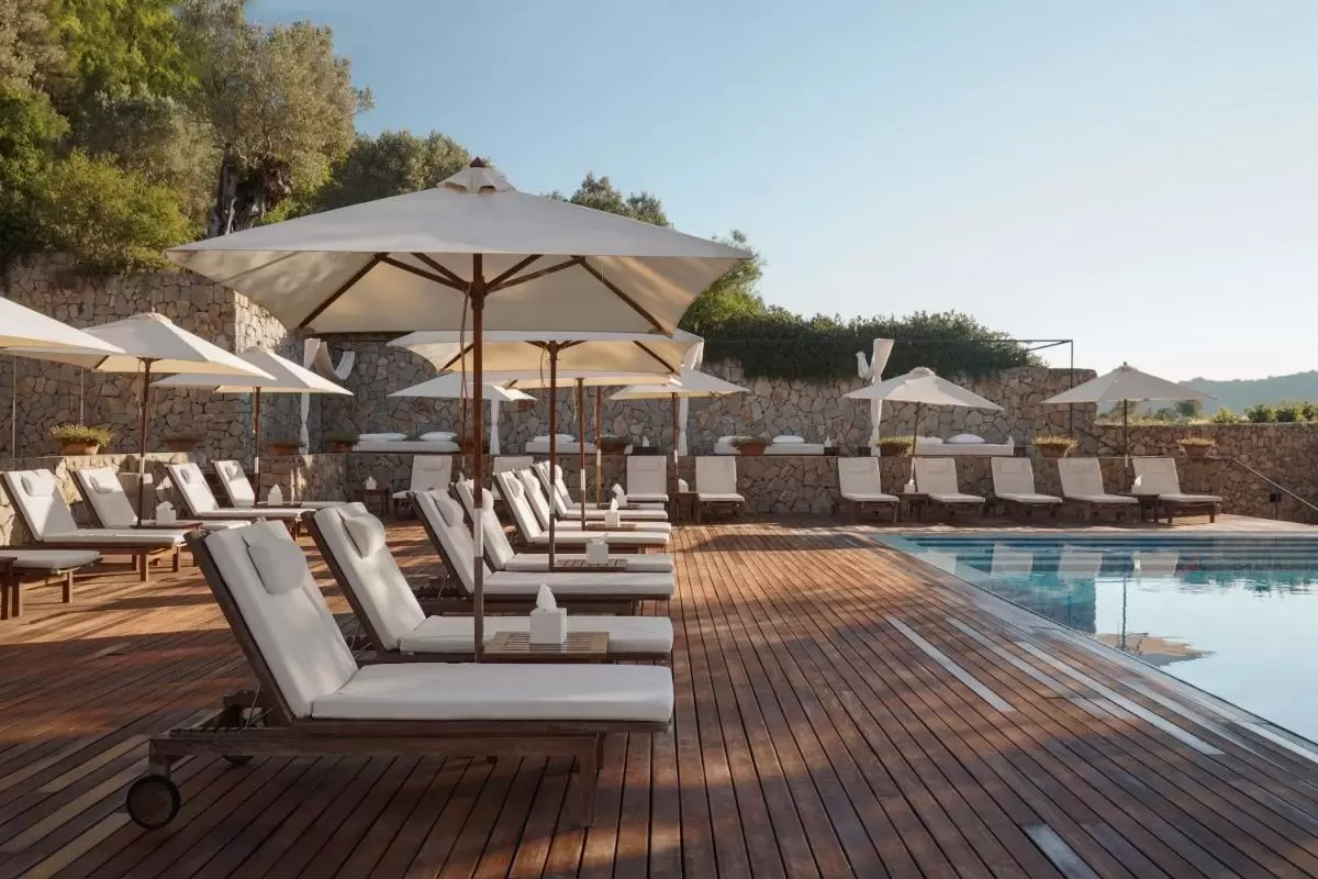 Área de piscina al aire libre con deck de madera, tumbonas con cojines blancos y sombrillas en Mallorca