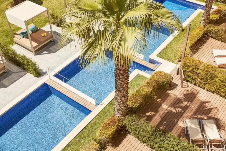Área exterior con piscina dividida en secciones, palmera central y zona de descanso con tumbonas y cama balinesa en Mallorca