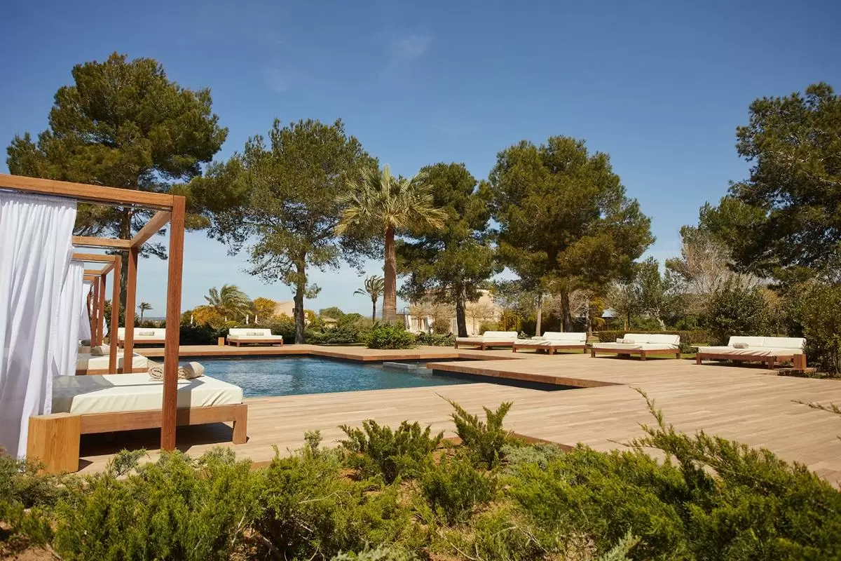 Área exterior con piscina rectangular, deck de madera y camas camastro con pérgolas y cortinas en Mallorca