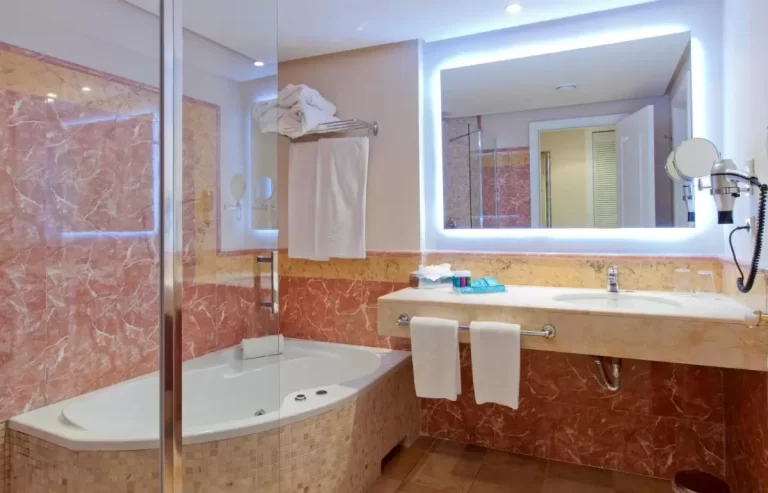 Baño con acabados en mármol rojizo y beige, bañera empotrada y lavabo con espejo iluminado en Mallorca