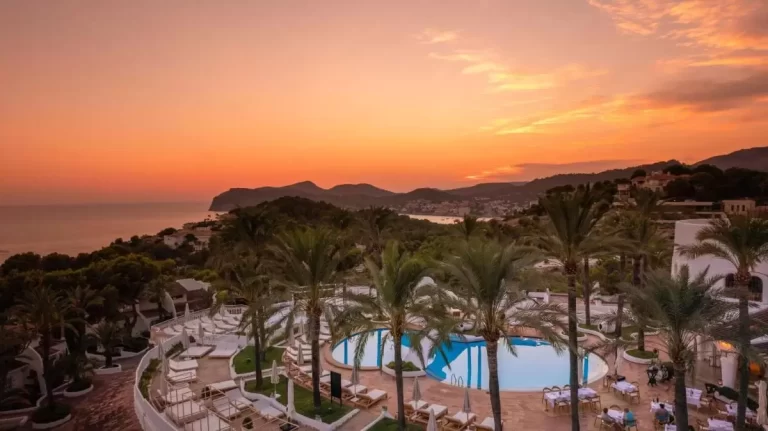 Escena al aire libre en Mallorca con piscina irregular, palmeras y vistas al mar durante el atardecer