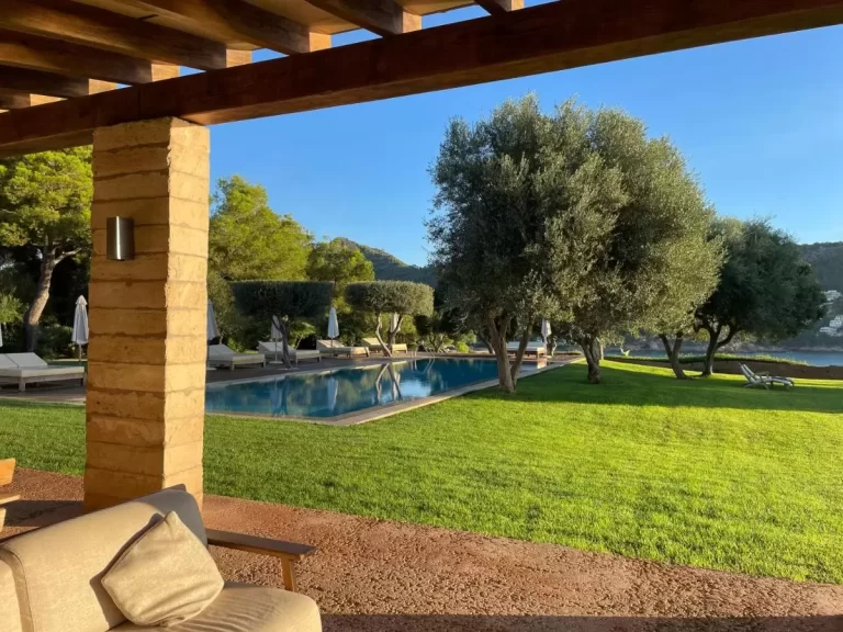 Espacio exterior con piscina rectangular, área de césped, estructura con columnas de piedra y vistas a colinas y cuerpo de agua en Mallorca