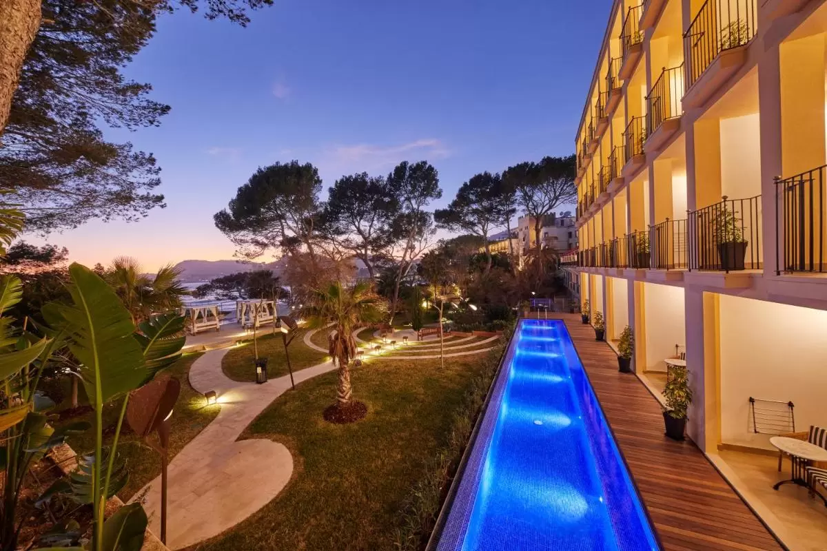 Espacio exterior en Mallorca con piscina iluminada, jardín con palmeras y pérgolas junto al mar al atardecer