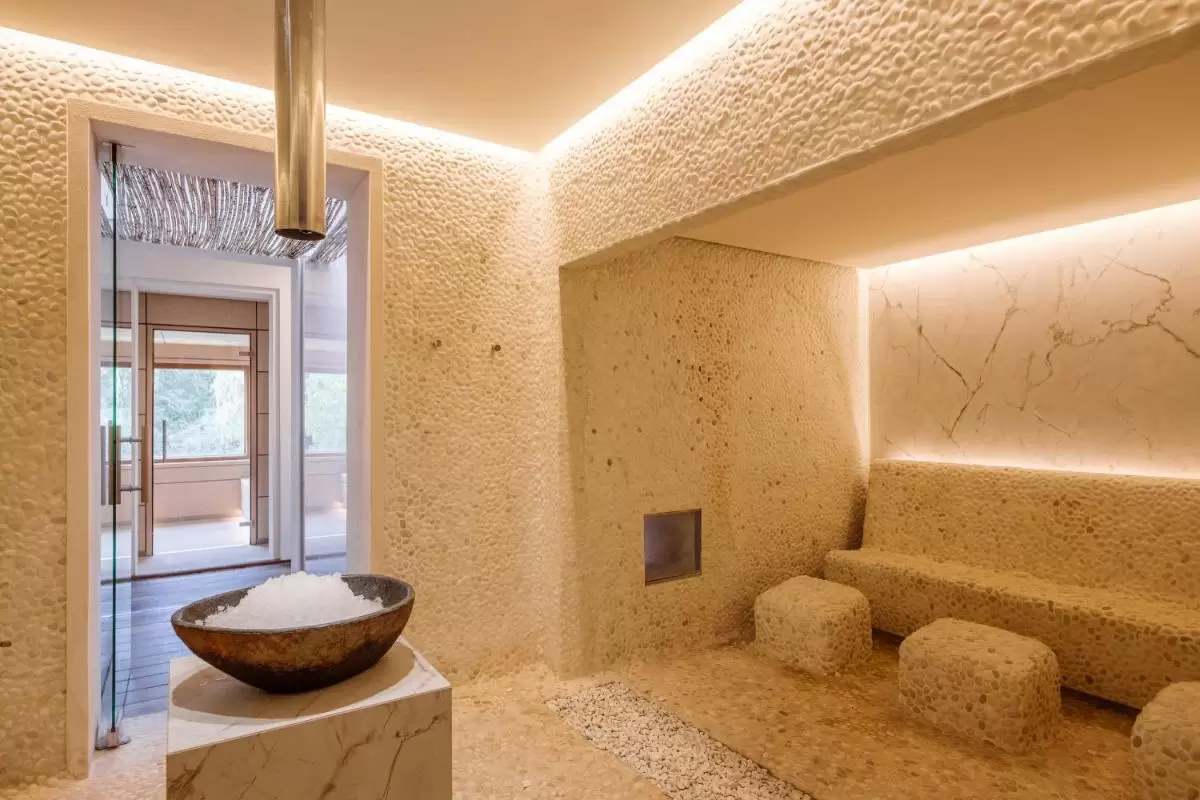 Espacio interior minimalista en Mallorca con revestimientos de piedra, banco y taburetes naturales junto a chimenea metálica y base de mármol