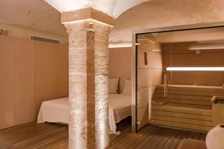 Espacio interior relajante con sauna de madera clara, columna de piedra iluminada y cama blanca en Mallorca