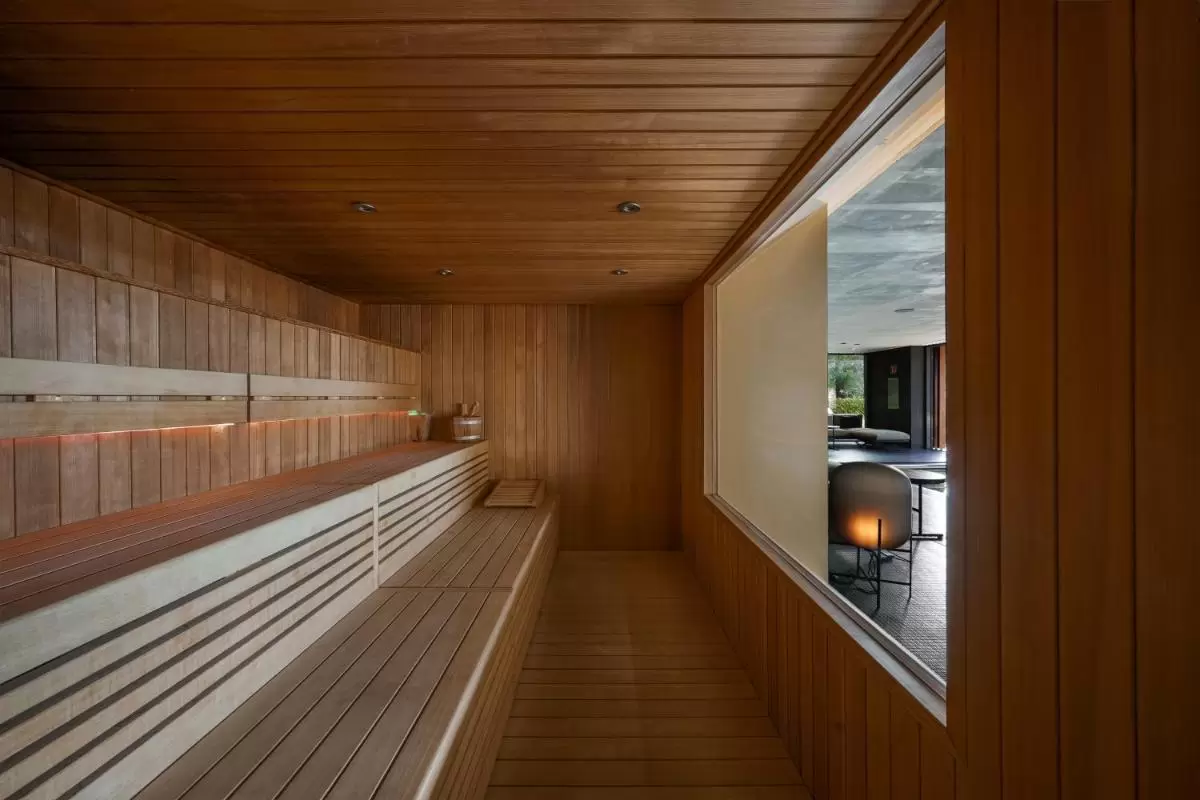 Interior de sauna de madera clara con bancos escalonados y ventana hacia sala moderna en Mallorca