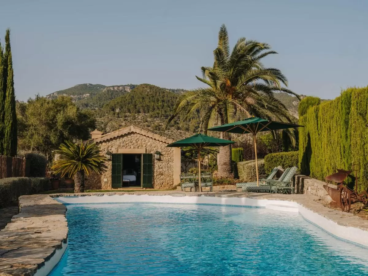 Piscina al aire libre con borde de piedra y zona de descanso con sombrillas verdes en entorno natural de Mallorca