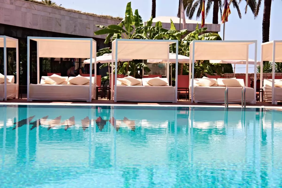 Piscina al aire libre con camas daybed blancas, toldos, plantas tropicales y banderas en Mallorca