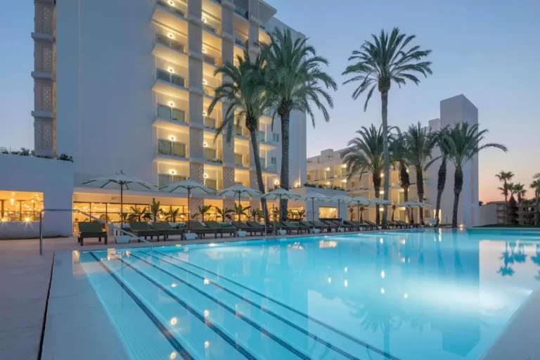 Piscina al aire libre con iluminación interior, tumbonas y palmeras en entorno moderno al atardecer en Mallorca