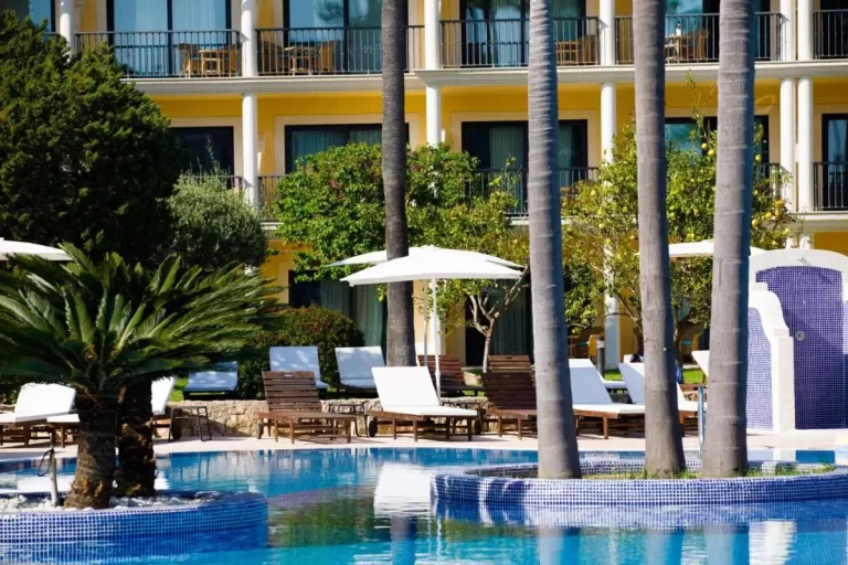 Piscina al aire libre con isla de palmeras, tumbonas de madera y sombrillas blancas en Mallorca