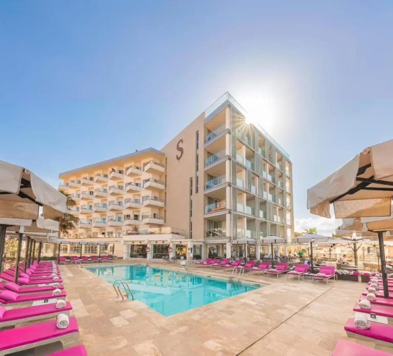 Piscina al aire libre con tumbonas rosas y sombrillas blancas en entorno moderno con edificio de varios pisos en Mallorca