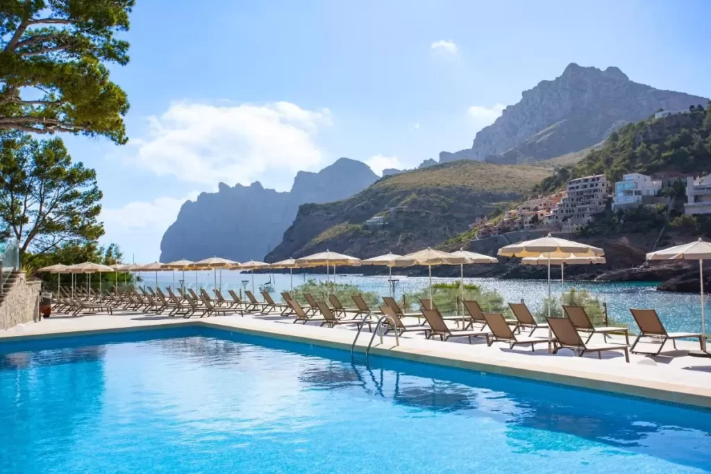 Piscina al aire libre con tumbonas y sombrillas frente al mar y montañas en Mallorca