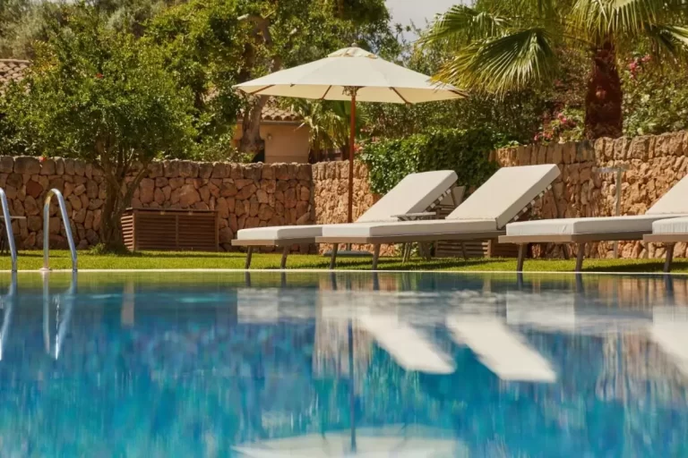 Piscina al aire libre en Mallorca con tumbonas, sombrilla blanca y entorno natural con árboles y muro de piedra