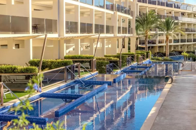 Piscina alargada con azulejos azules, barandales de vidrio y áreas de descanso con césped artificial en Mallorca