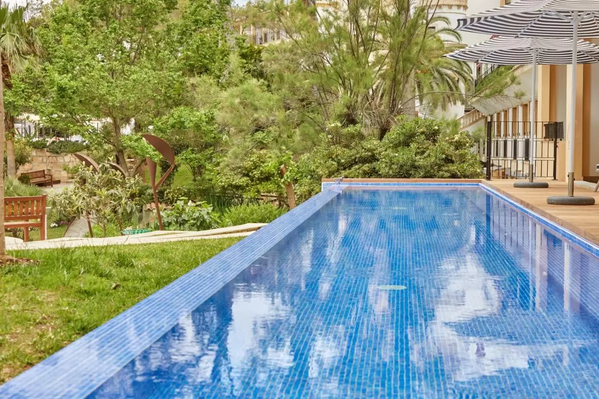 Piscina alargada con azulejos azules, deck de madera y jardín con esculturas en Mallorca