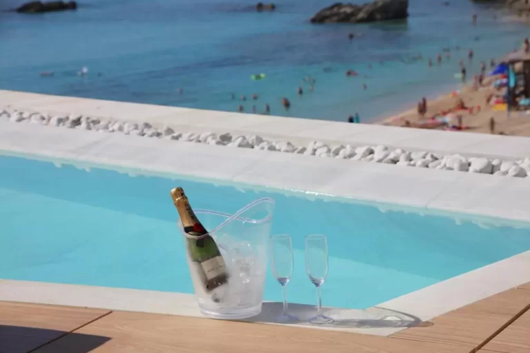 Piscina de borde infinito con champaña y copas frente a playa de arena clara y mar turquesa en Mallorca
