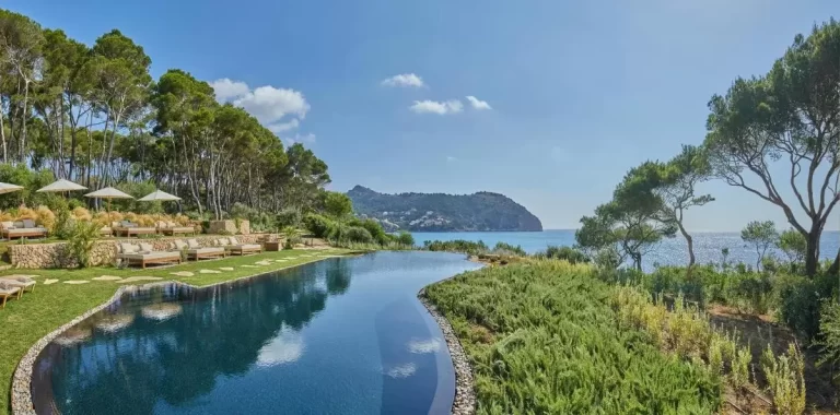 Piscina de borde infinito con forma curva en entorno natural junto al mar en Mallorca con tumbonas y vegetación mediterránea