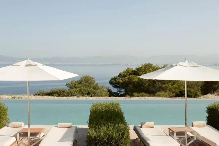 Piscina de borde infinito con tumbonas y sombrillas en entorno natural frente a costa y montañas en Mallorca