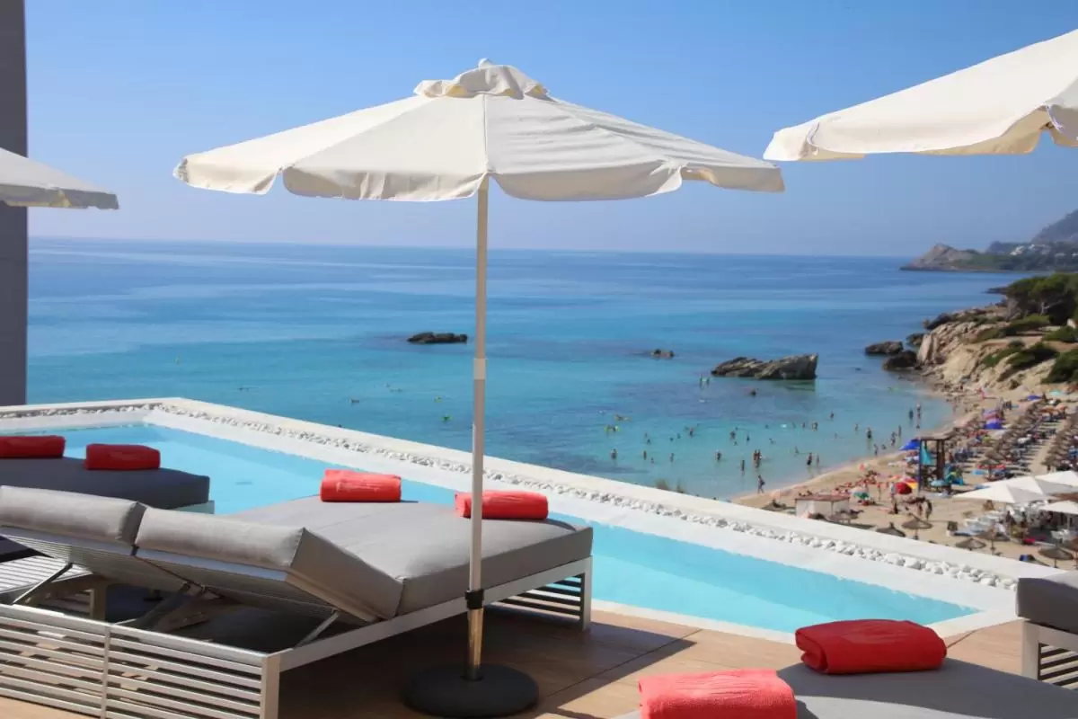 Piscina elevada con borde de piedras blancas y tumbonas bajo sombrillas frente a playa de Mallorca