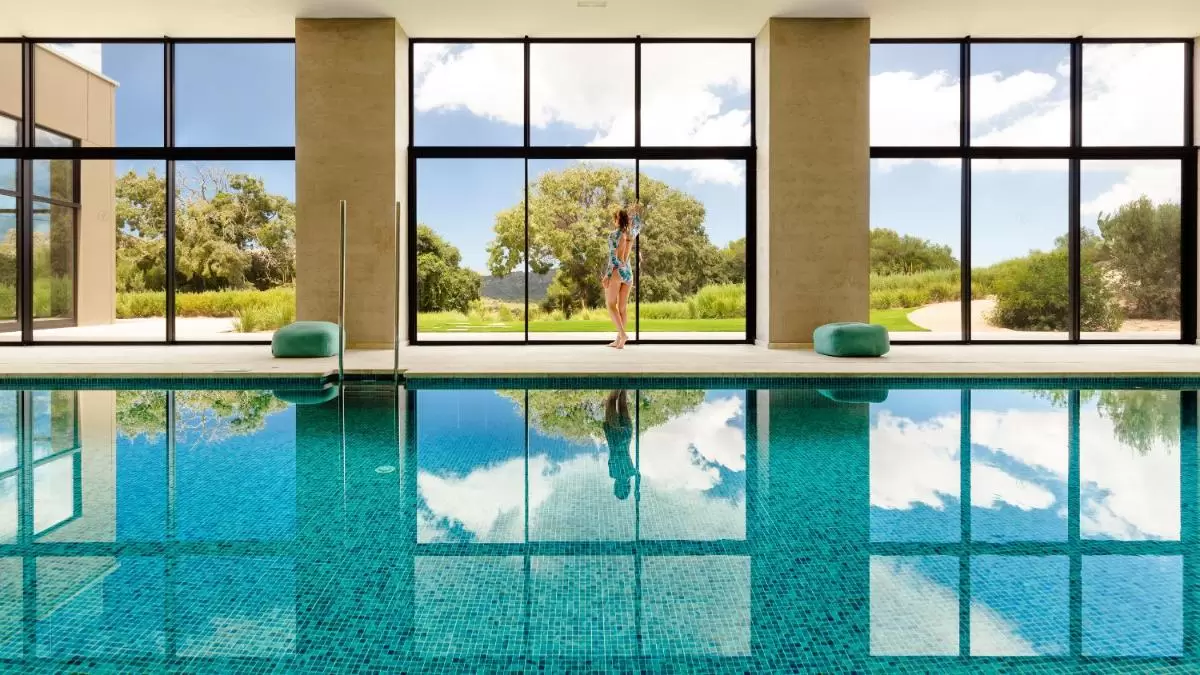 Piscina interior con azulejos azules y grandes ventanales que integran el espacio con paisaje natural en Mallorca