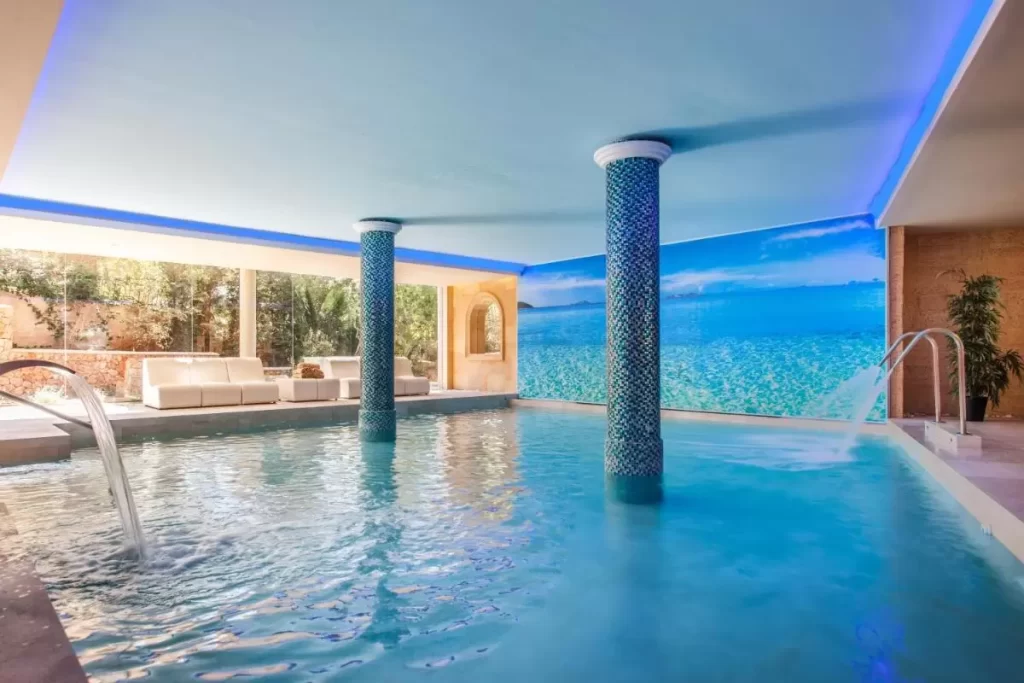 Piscina interior con columnas de mosaicos, iluminación azul y zona de descanso con vistas a jardín en Mallorca