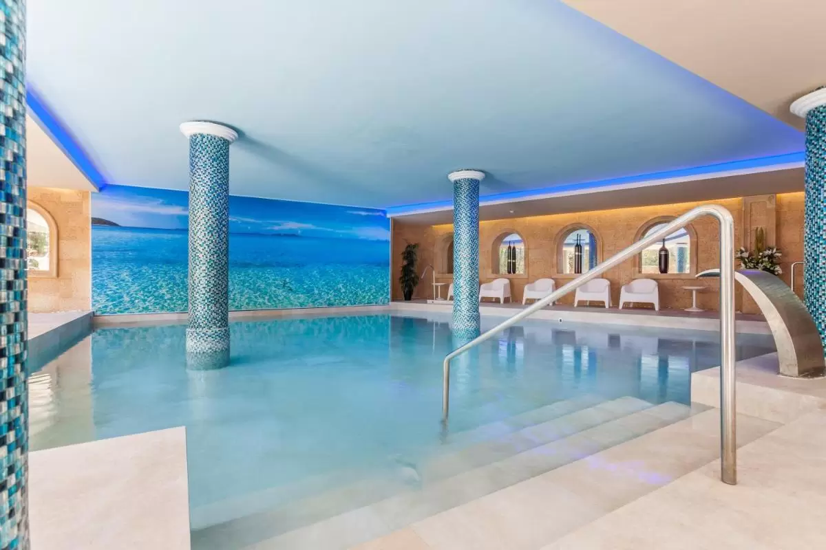 Piscina interior con mosaicos azules y verdes, iluminación LED azul y zona de descanso con sillas blancas en Mallorca