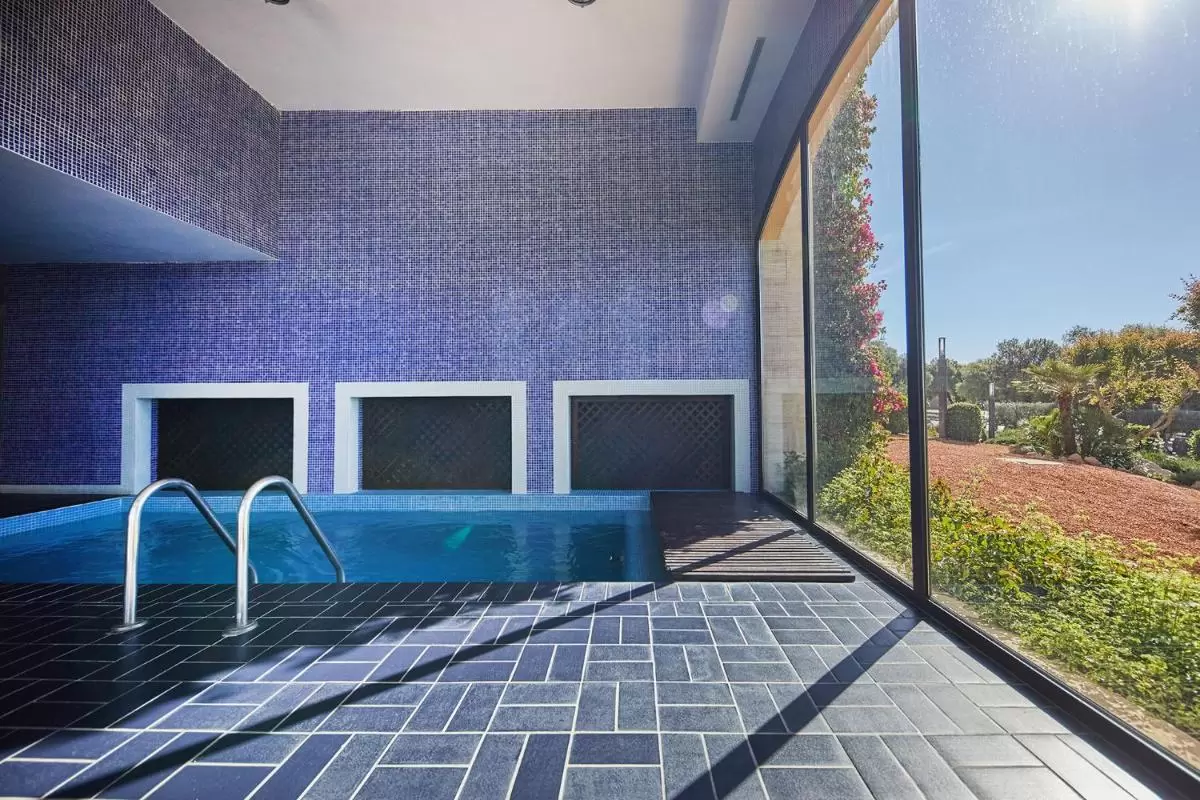 Piscina interior moderna con mosaicos azules y gran ventana hacia jardín en Mallorca