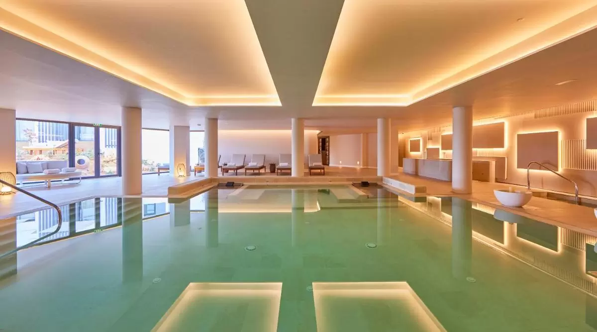 Piscina interior moderna en Mallorca con iluminación cálida, ventanales amplios y área de descanso con tumbonas blancas