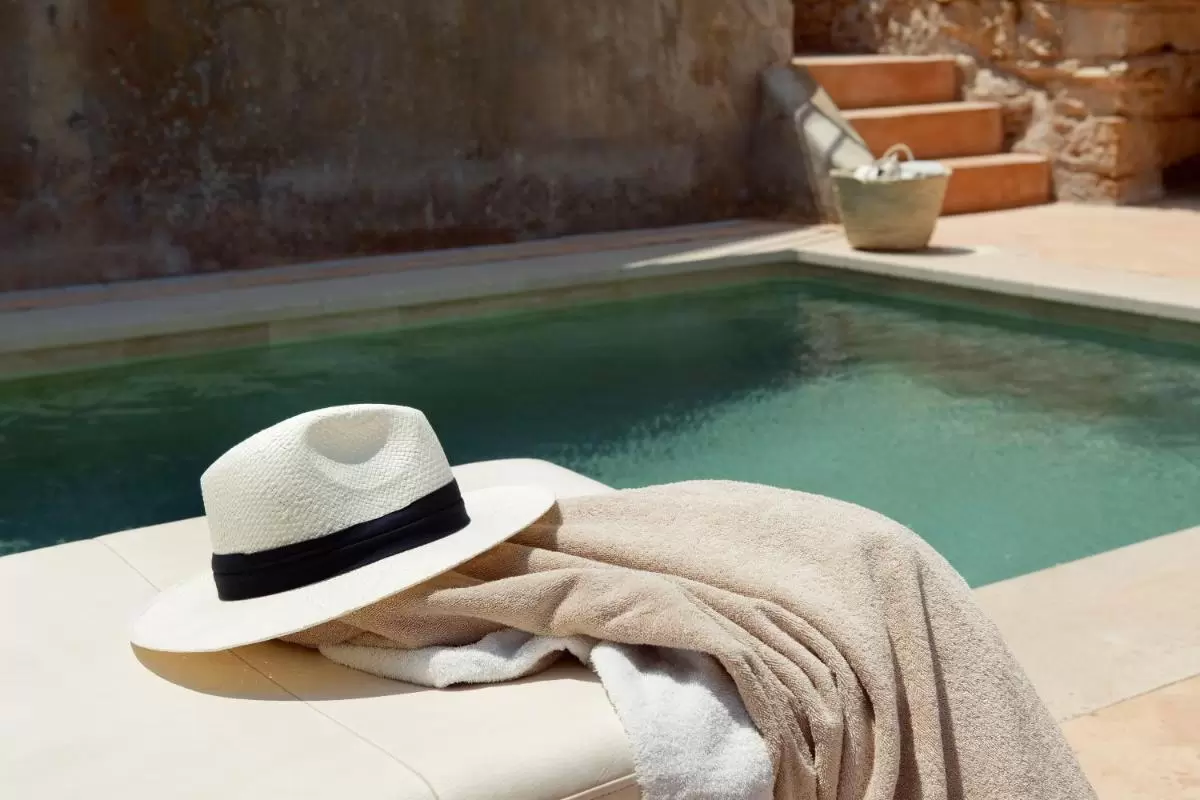 Piscina pequeña con agua verde azulado, borde de piedra beige y detalles de sombrero y toalla en Mallorca