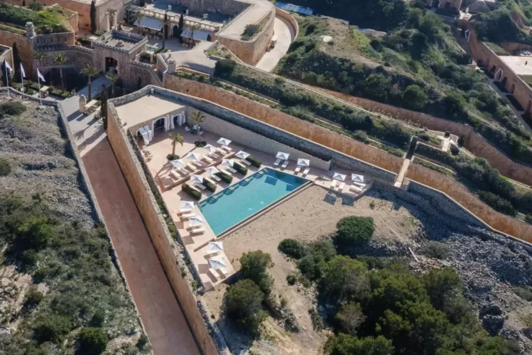 Vista aérea de piscina trapezoidal en estructura fortificada con muros de piedra y palmeras en Mallorca